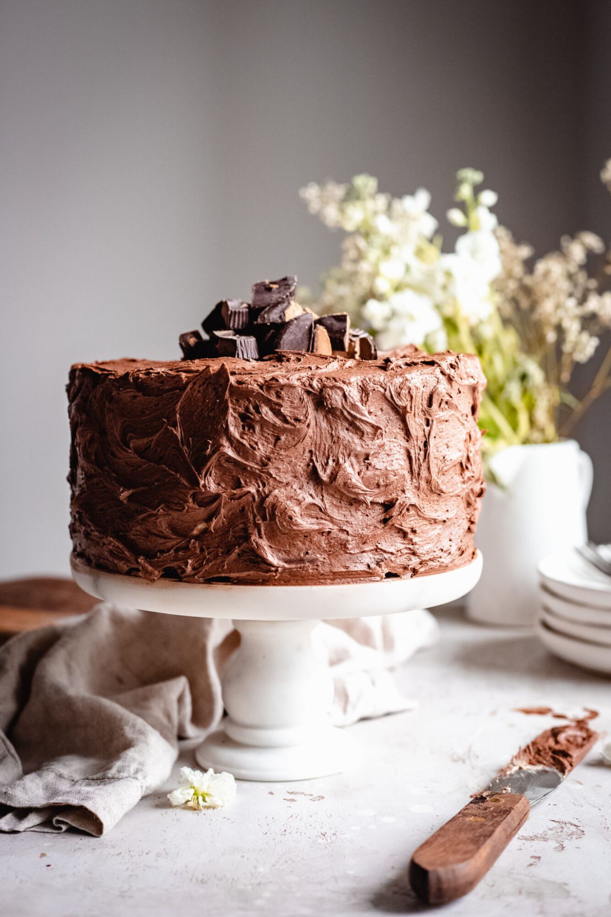 an iced chocolate cake sits on a white cake stand topped with broken peanut butter cups. a palette knife lies in front and flowers in a vase behind
