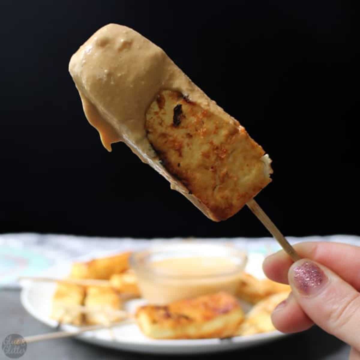 a hand holds a tofu skewer dipped into satay sauce. blurred in teh background is a plate full of tofu skewers