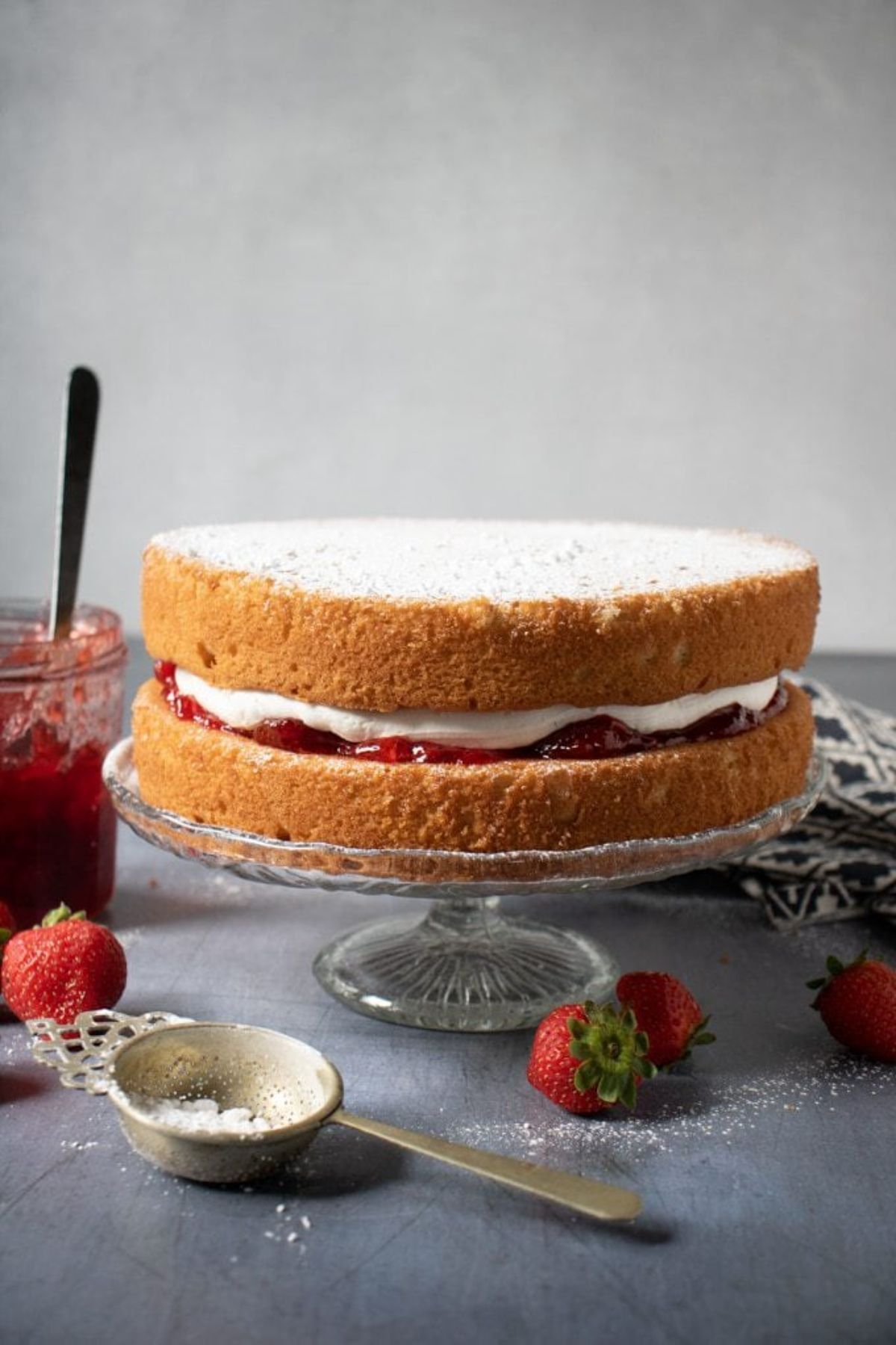 on a glass cake stand is a victoria sponge cake, dusted with icing sugar and filled with cream and straberery jam