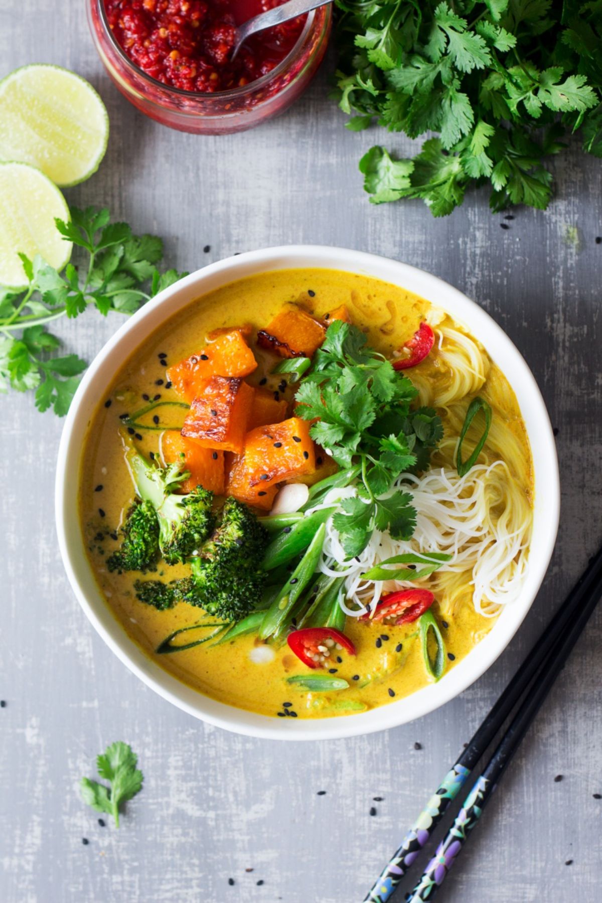 a white bowl full of khao soi. Fresh herbs and sliced limes sit to the side next to some chopsticks