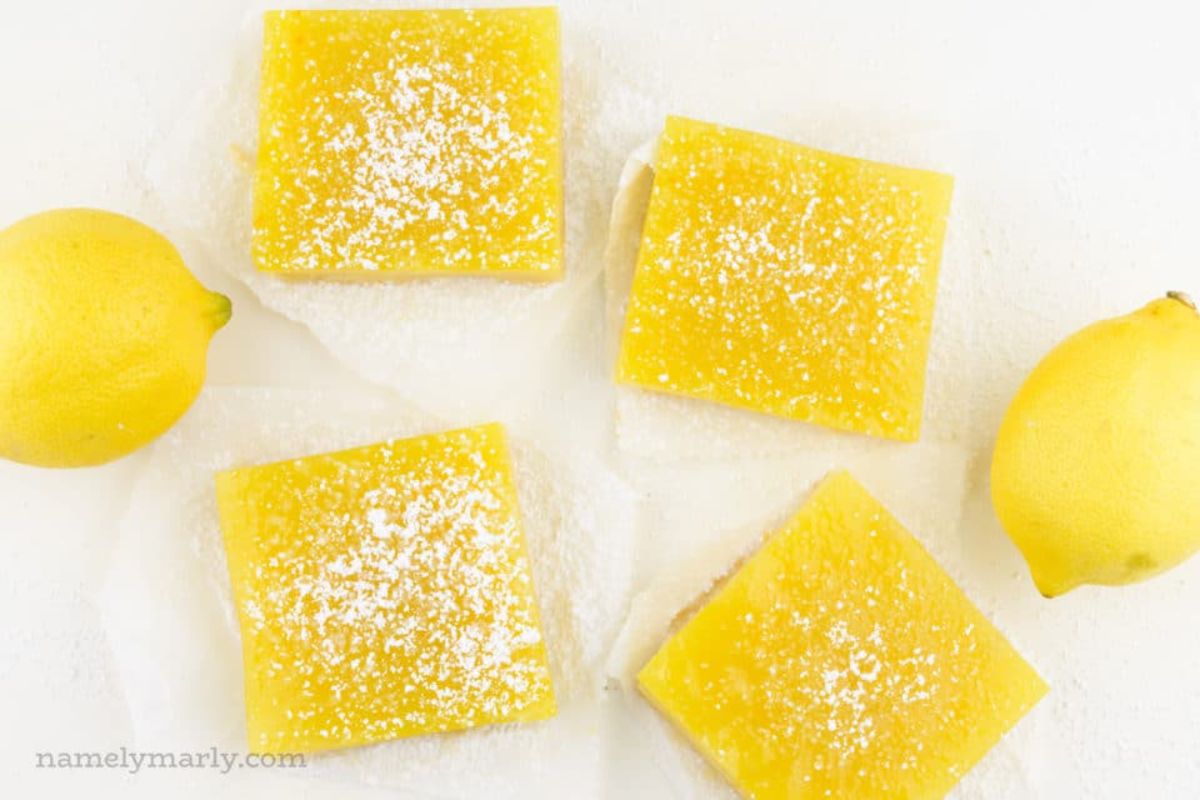 4 lemon bars sit on baking paper dusted with icing sugar next to 2 lemons