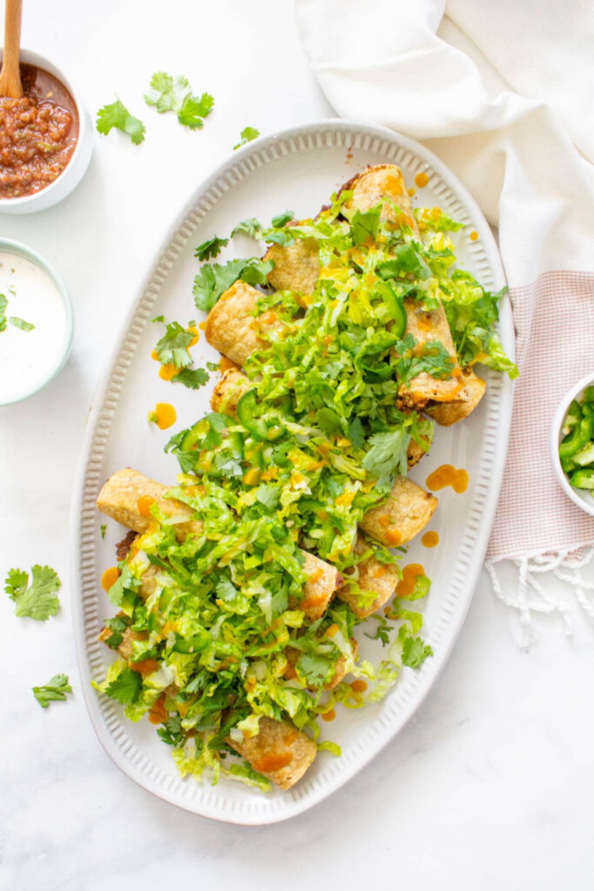 a top view of an oval dish filled with rolled tacos scattered with jalapeno slices and chopped lettuce