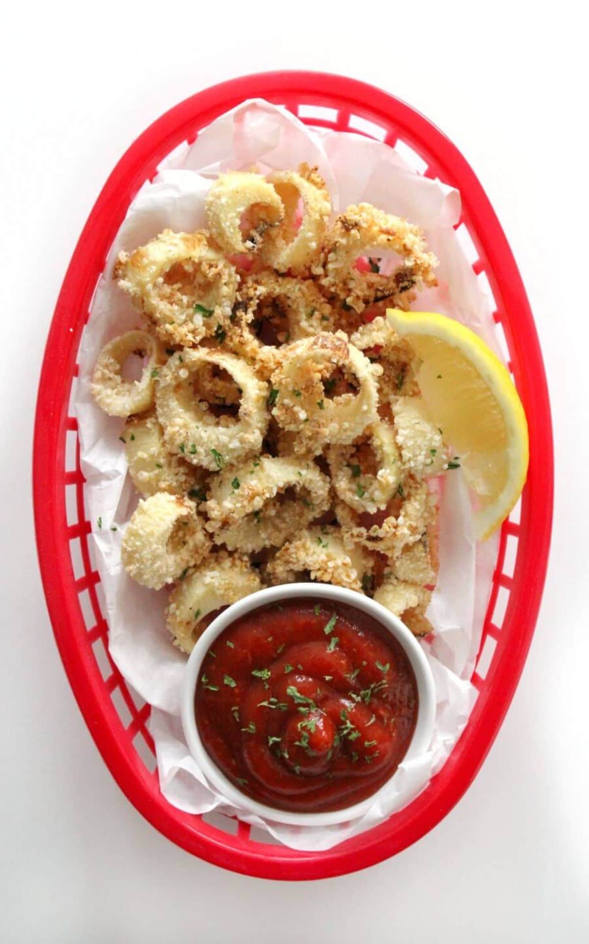 a top view of a red woven basket filled with vegan calamari rings next to a lemon slice and a bowl of red sauce