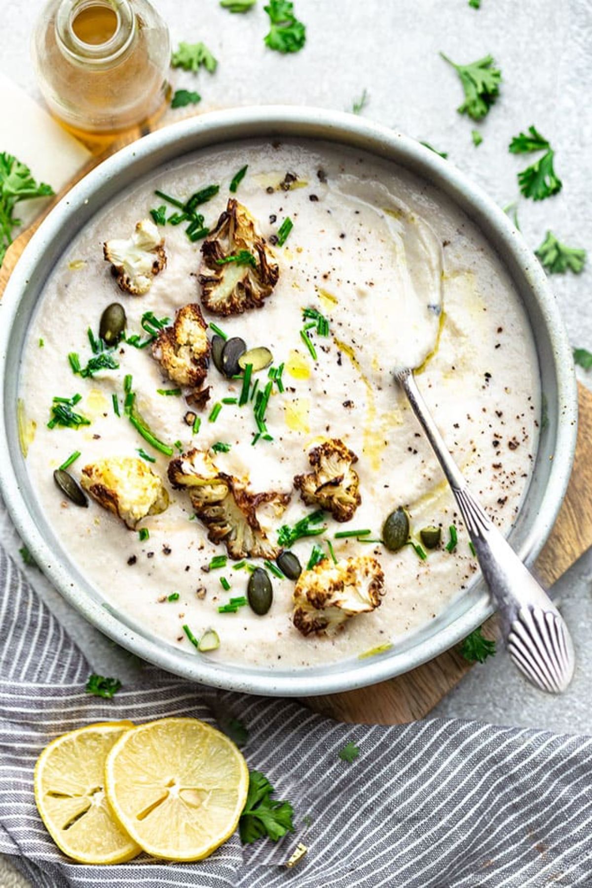 a bowl full of creamy cauliflower soup, topped with scattered herbs and pumpkin seeeds. Sliced lemons and herbs are on the side