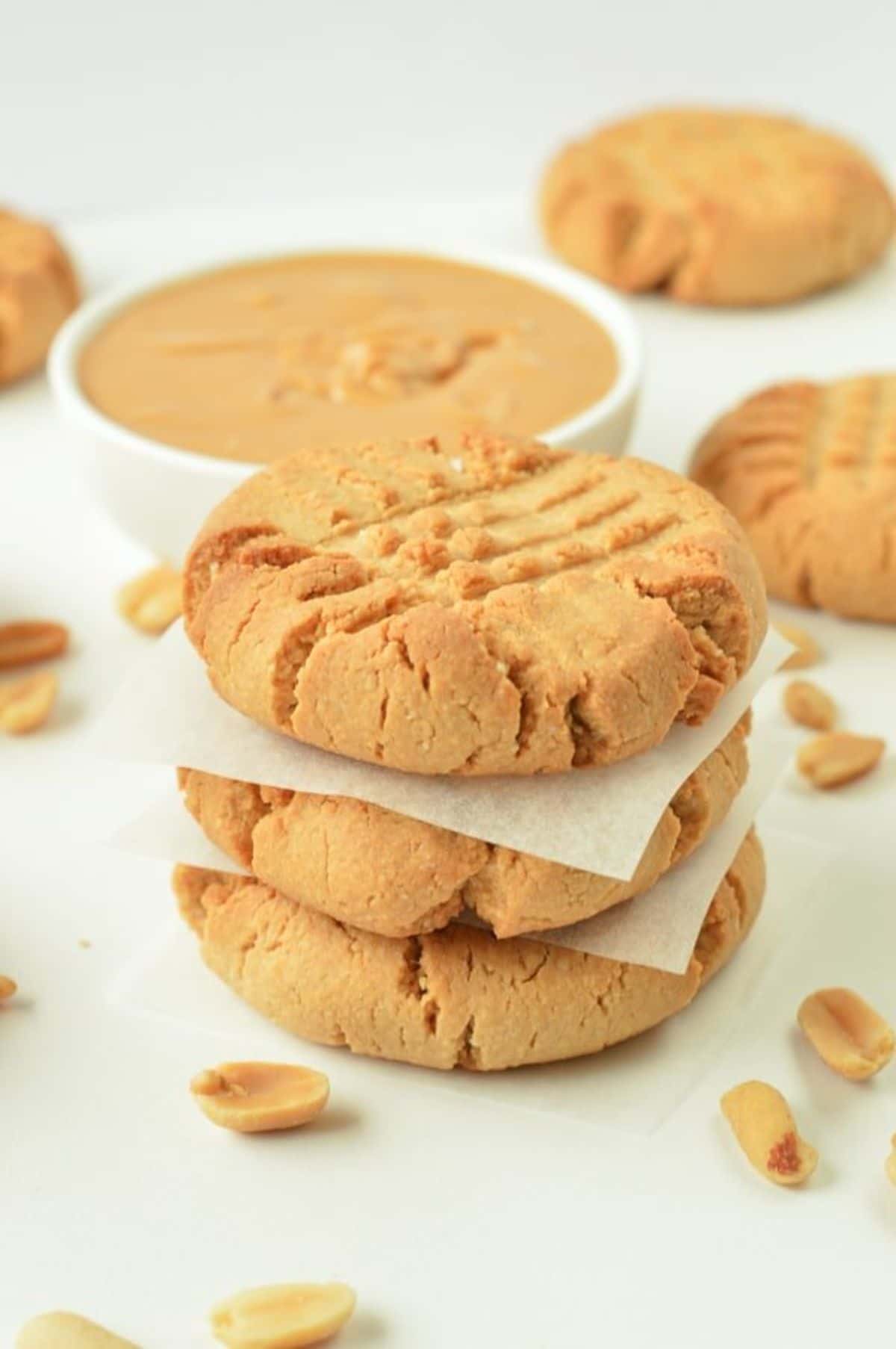 a pile of peanut butter cookies with a bowl of peanut butter and some scattered cookies behind it