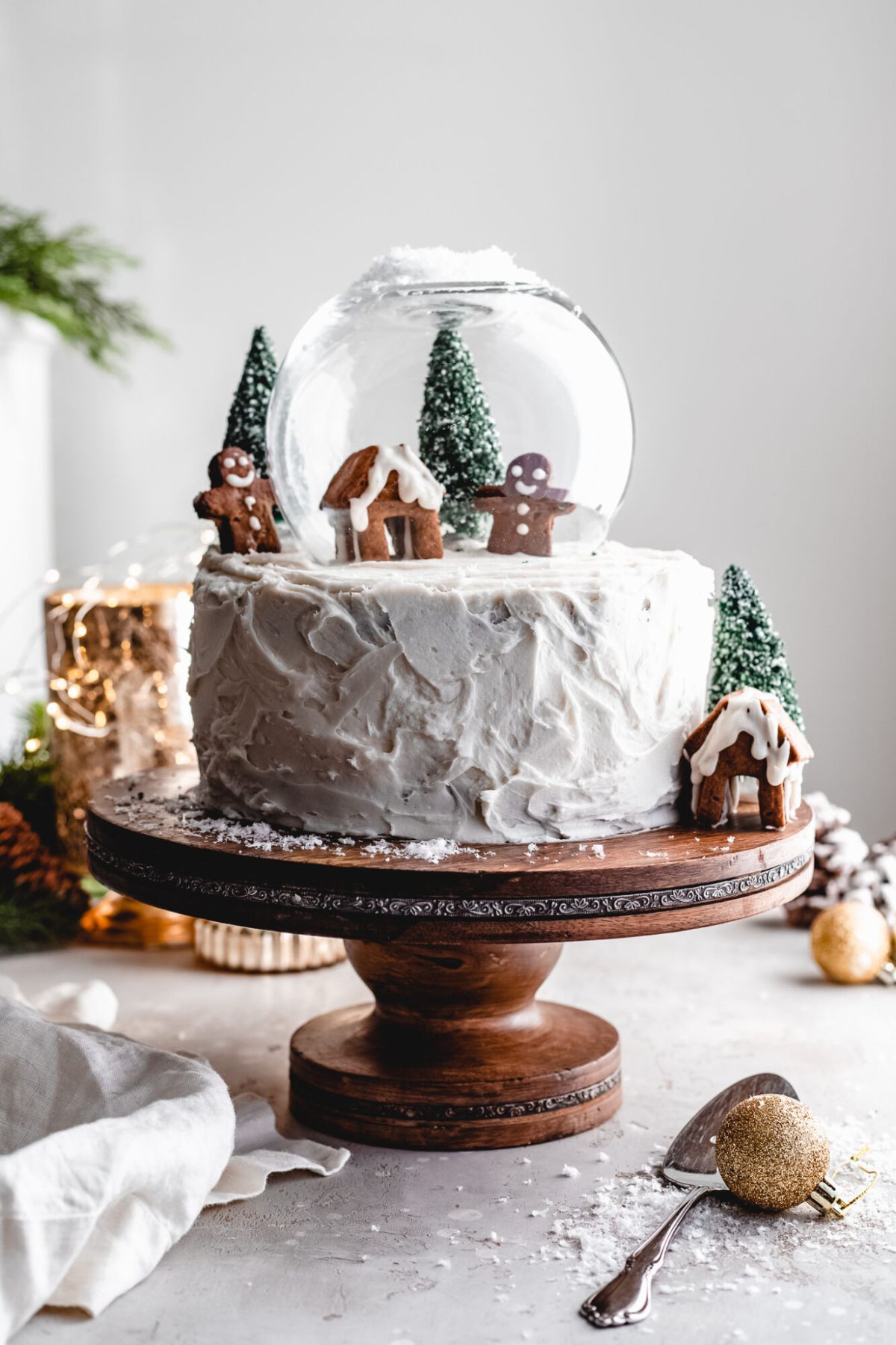 on a wooden cakes stand is a vegan ginger cake decorated to look like a christmas scene. gingerbread men, christmas trees and a gingerbread house sit under a snowglobe on top of the cake