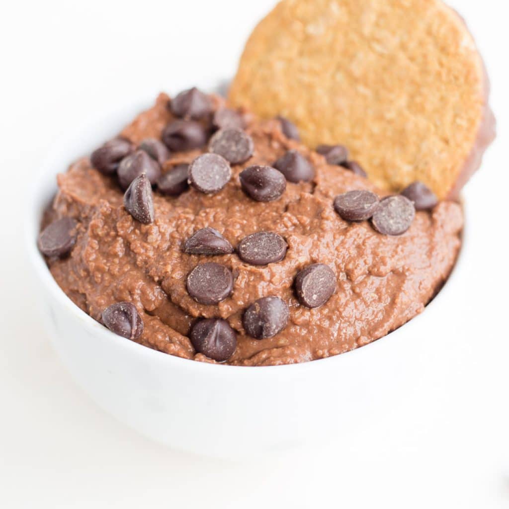 Brownie Batter Chocolate Hummus in small white bowl with chocolate chips and a cookie on top.