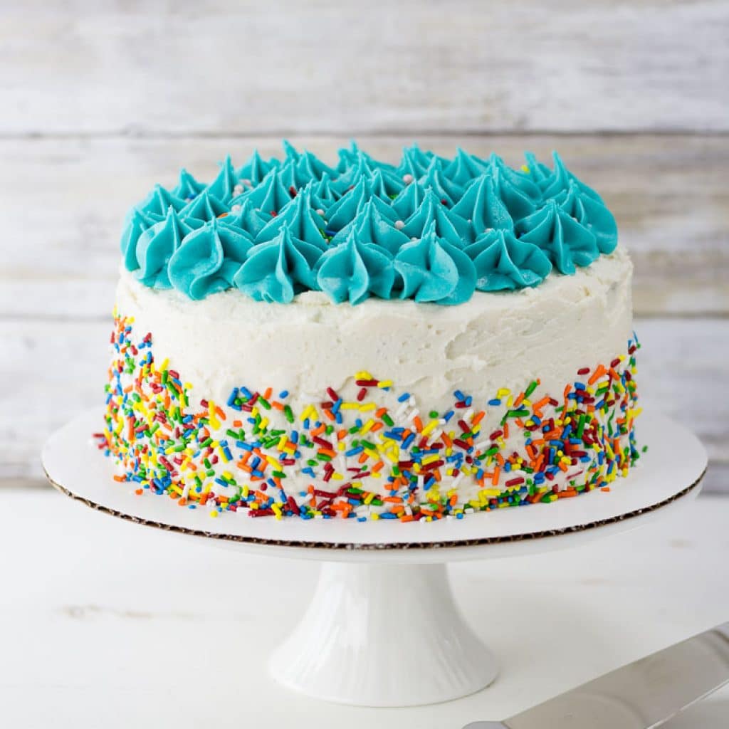 Gluten Free Birthday Cake with white and blue frosting and sprinkles on the side on a cake stand
