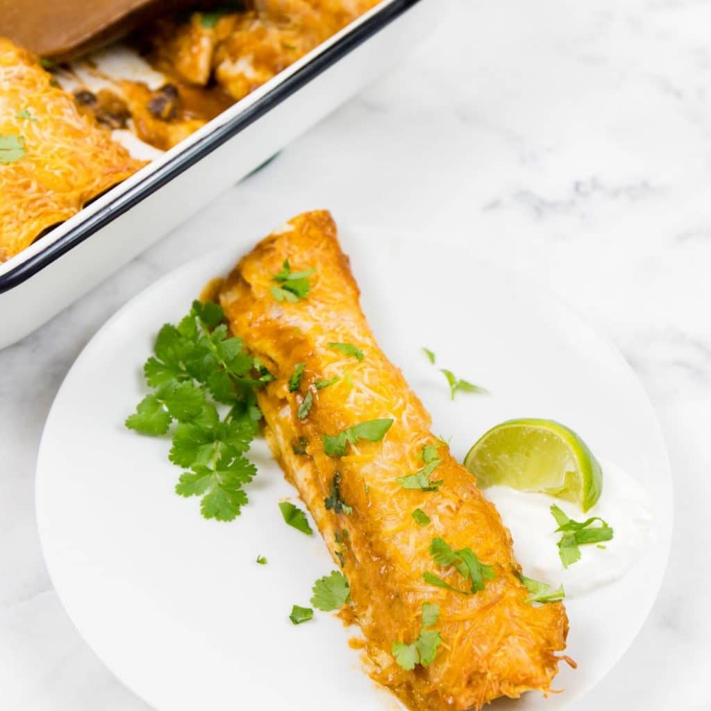 Black Bean Enchilada on a white plate with a slice of lime and a casserole dish of Black Bean Enchiladas in the background.