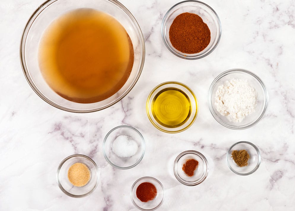 Ingredients you'll need for Homemade Red Enchilada Sauce in small bowls on a white surface