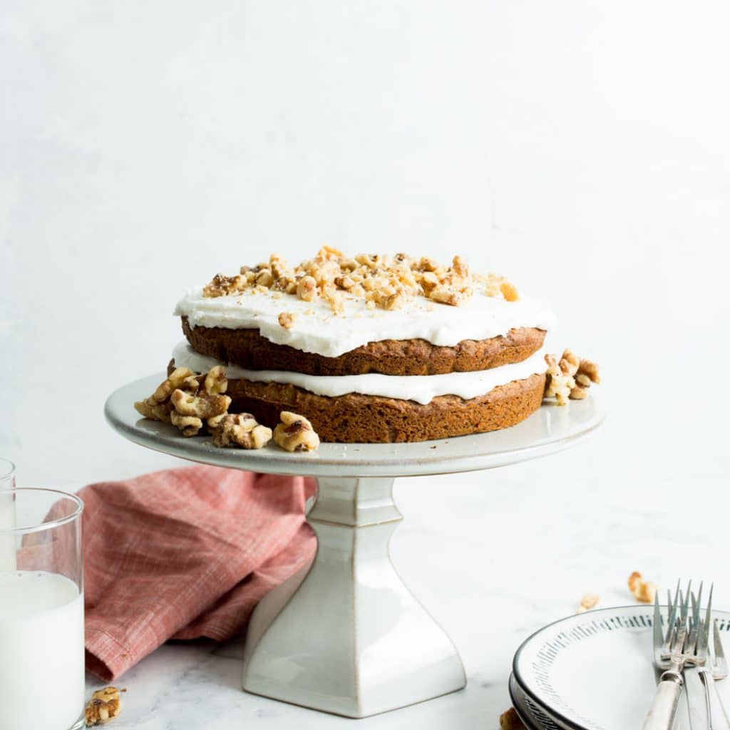 Vegan Carrot Cake on a white cake stand with two glasses of milk and a stack of plates.