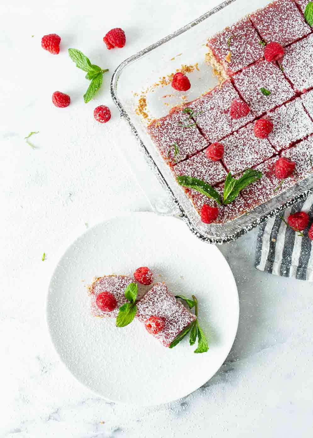 Overheat shot of pan of vegan raspberry bars and a small white plate in lower left corner with two bars all decorated with fresh raspberries