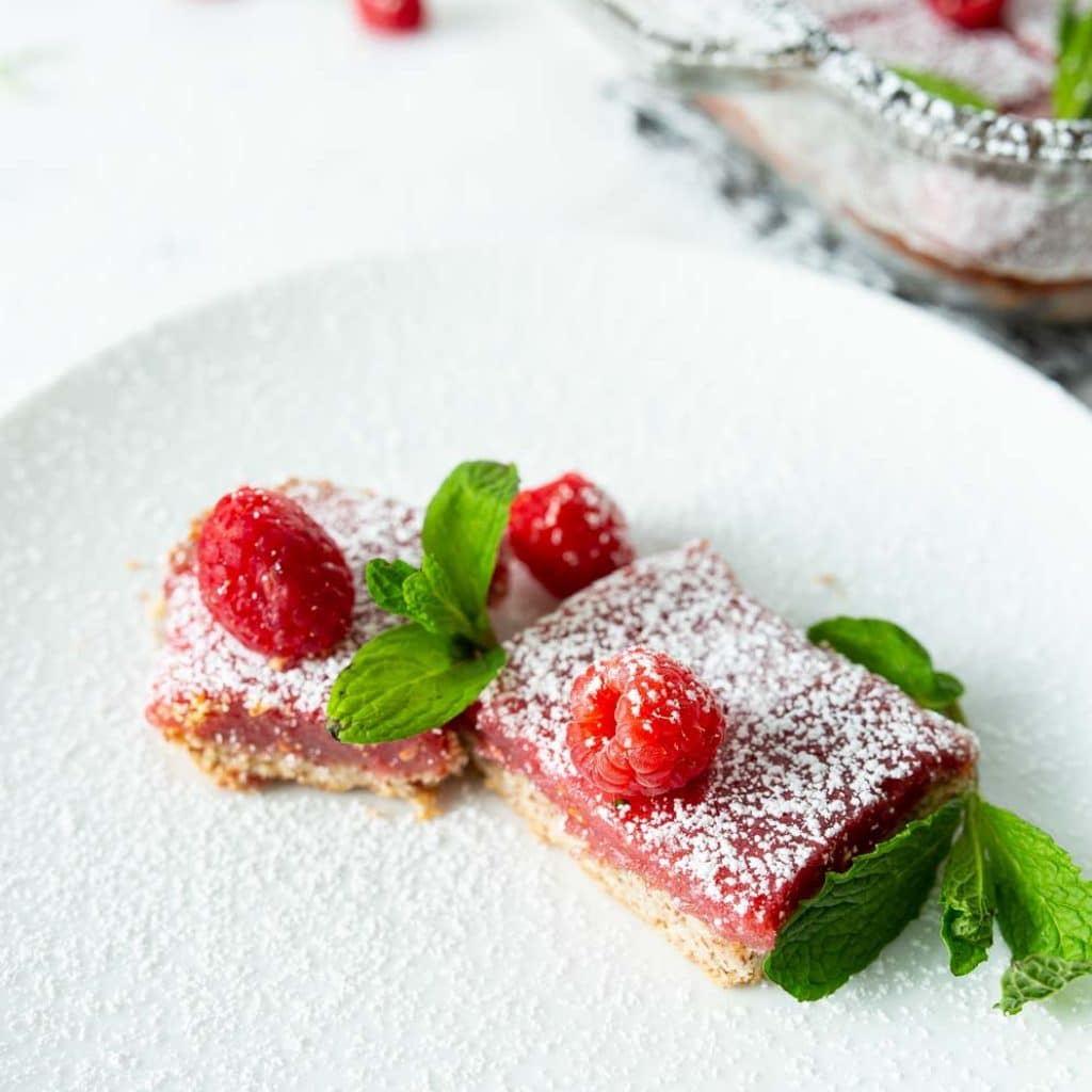 Two Vegan Raspberry Lemon Bars with fresh raspberries and mint leaves decorated around on a small plate
