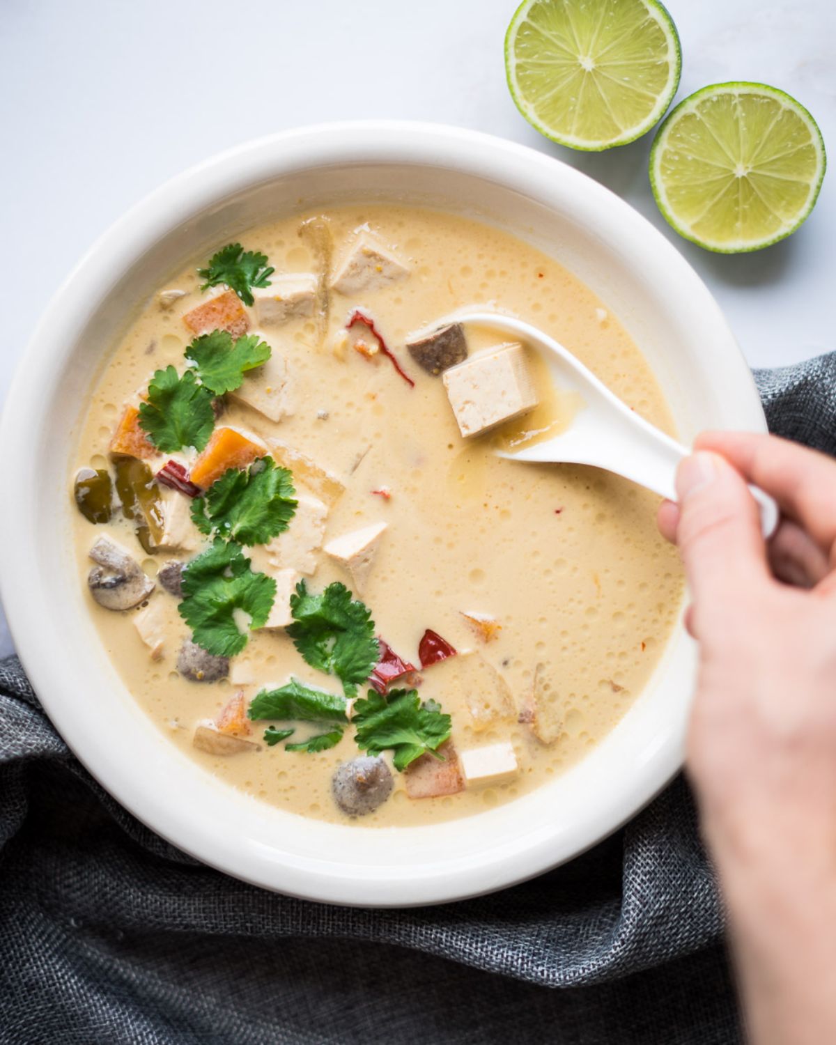 a white bowl with a china soup filled with tom kah, topped with herbs. halved limes sit to the right hand side