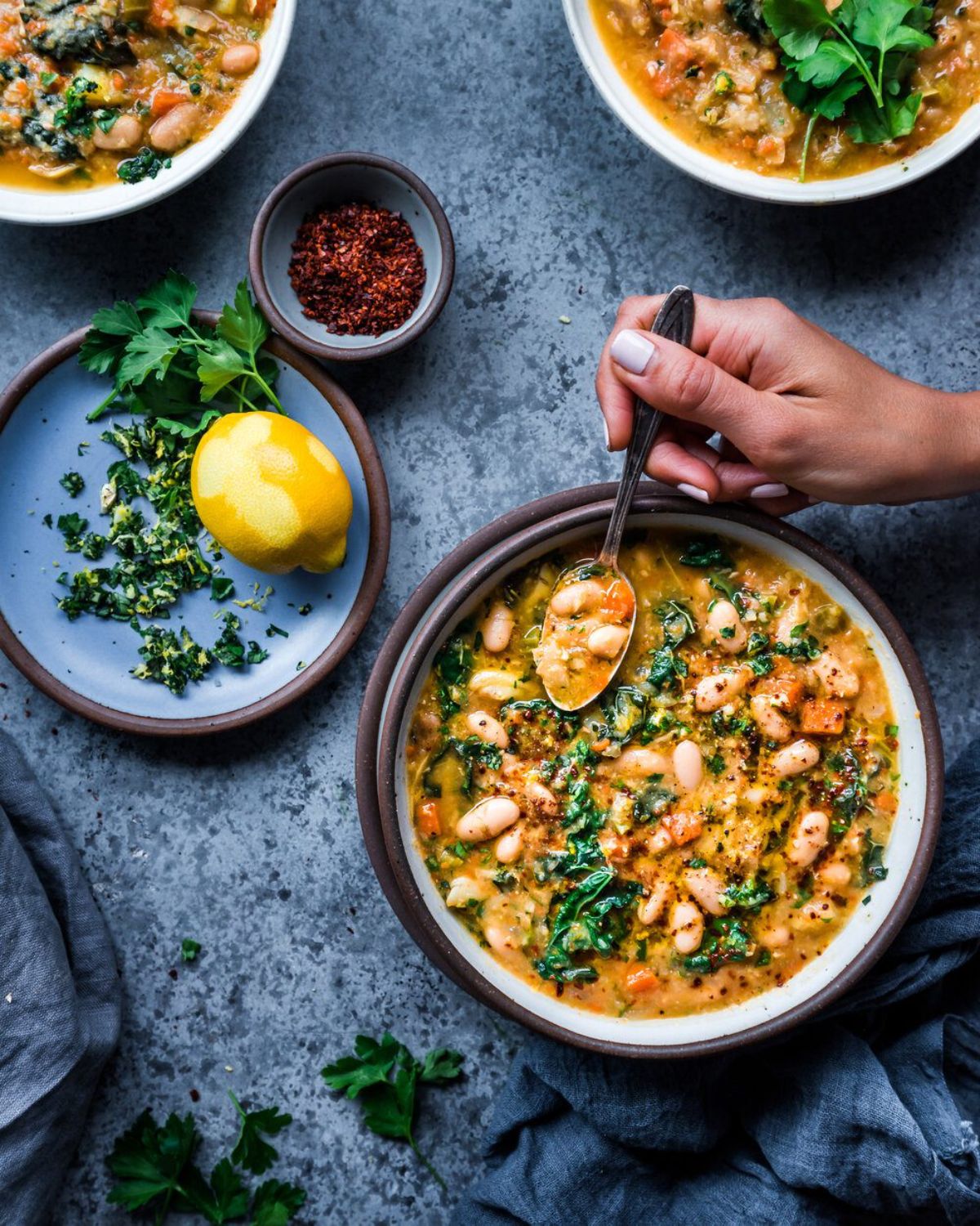 a hand dips a spoon into a white bean and kale soup bowl. a lemon and herbs sit to the side