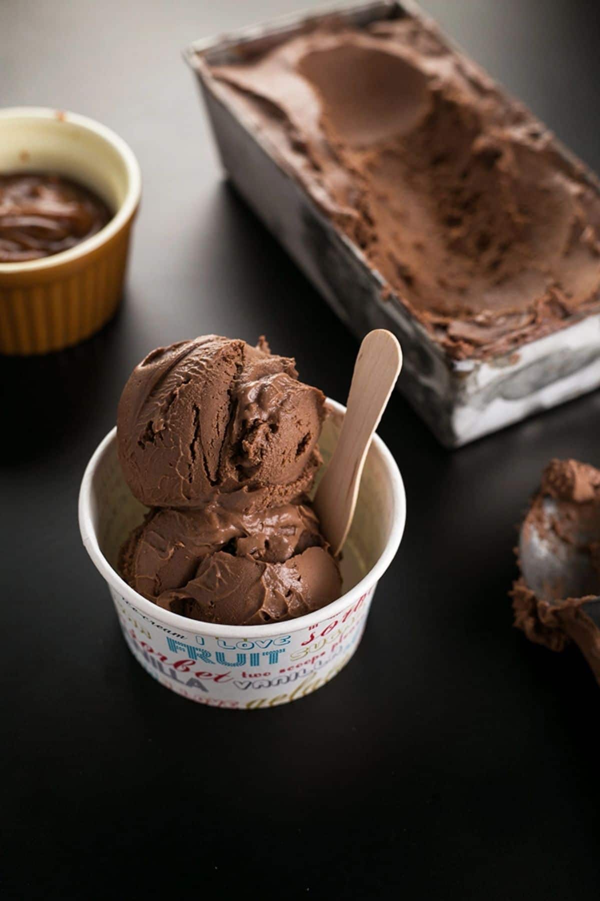 a paper cup filled with vegan chocolate ice cream with a wooden spoon sticking in it. in the background is a metal tray of ice cream
