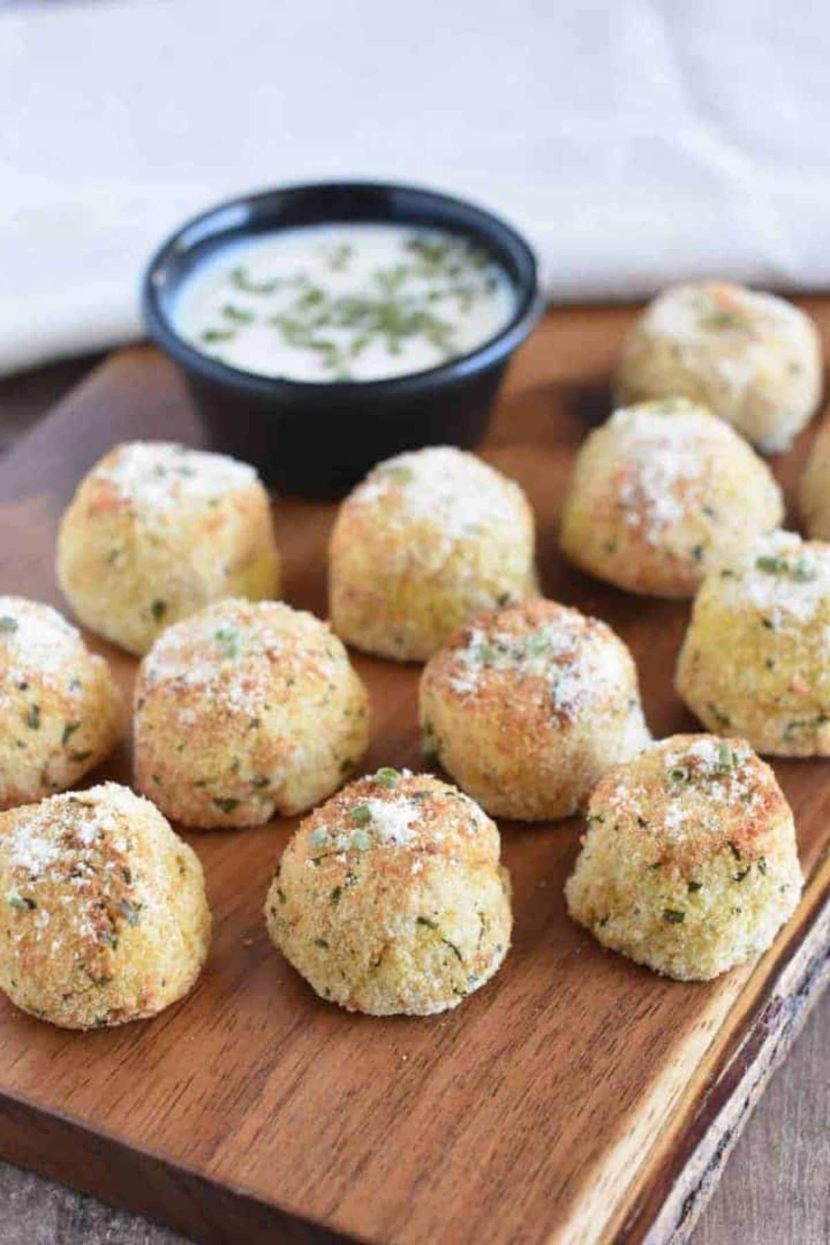 mashed potato balls sit on a wooden board next to a bowl of white dip scattered with herbs