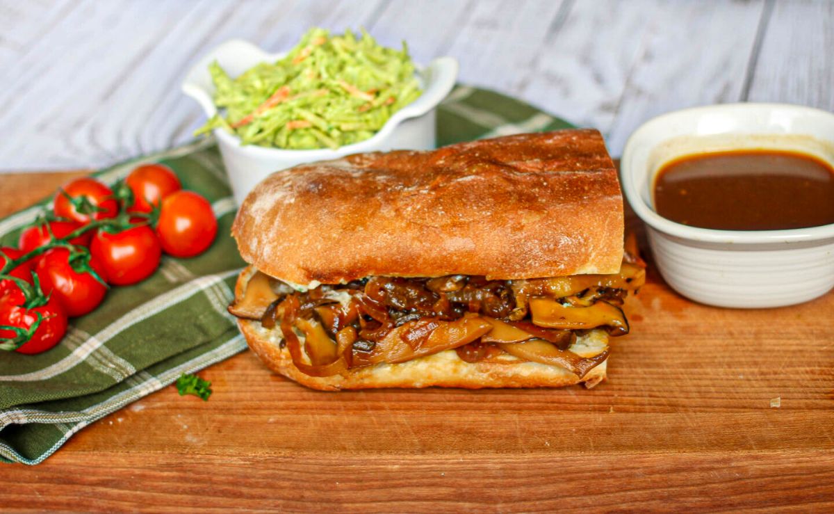 a baguette filled with vegan mushroom french dip sits on a board with a vine of tomatoes, shredded lettuce, and dip