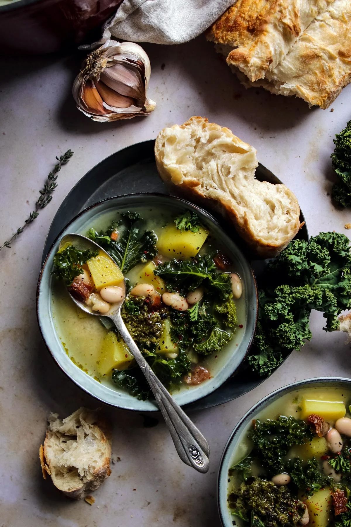 a bowl of zuppa toscana sits on a plate with a slice of bread and a pile of kale. A bulb of garlic, a loaf of bread and herbs sit to the side