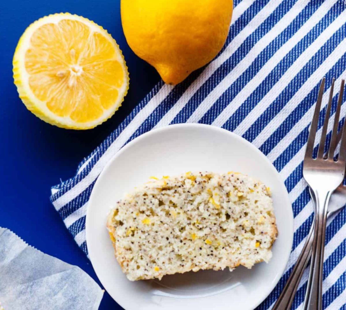 on a striped blue cloth is a white plate with a slice of lemon and poppy seed cake on it. A fork sits next to it and 2 lemons behind