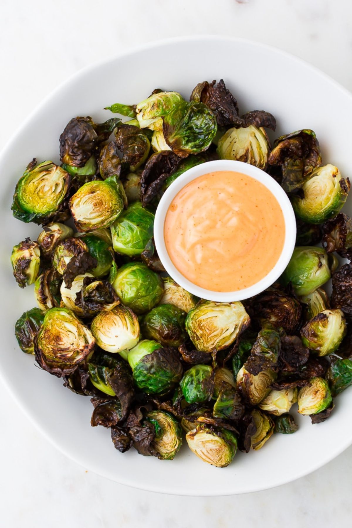 a top view of a white bowl full of air fried brussels sprouts with a bowl of orange dip in the middle