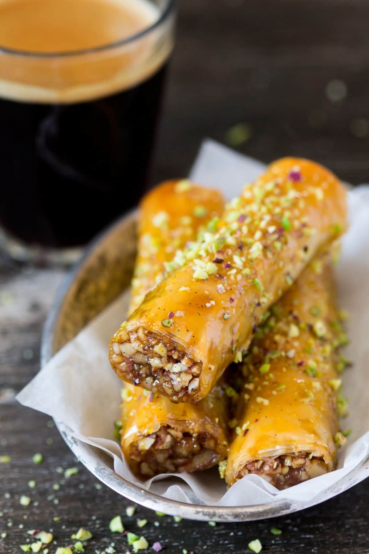 3 vegan baklava cigarssit on a plate with a black coffee behind it