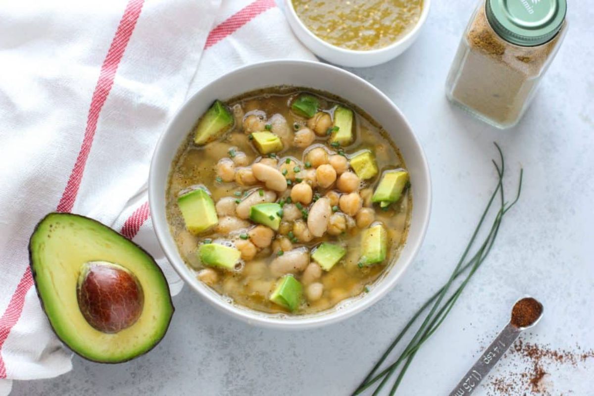 a white bowl full of chili verde, next to half an avocado, herbs and spices