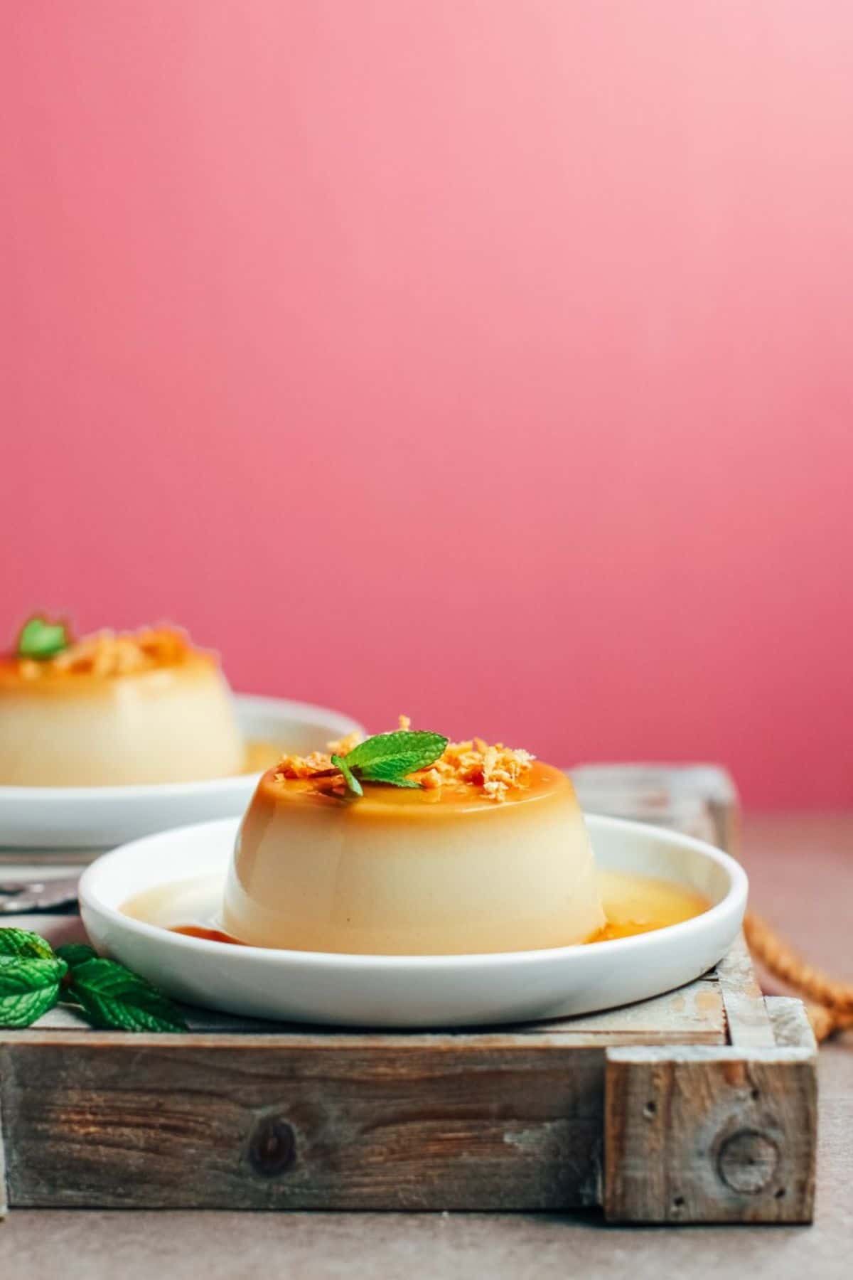 2 vegan creme caramels sit on white plates against a pink wall. They are topped with mint leaves