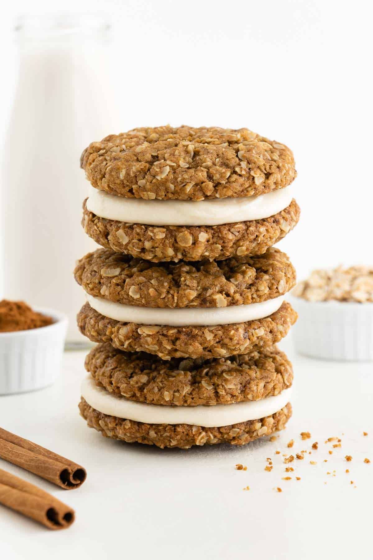 a pile of oatmeal cream pies are on a white surface surrounded by a bow of cinnamon, a bowl of oats, and 2 cinnamon sticks