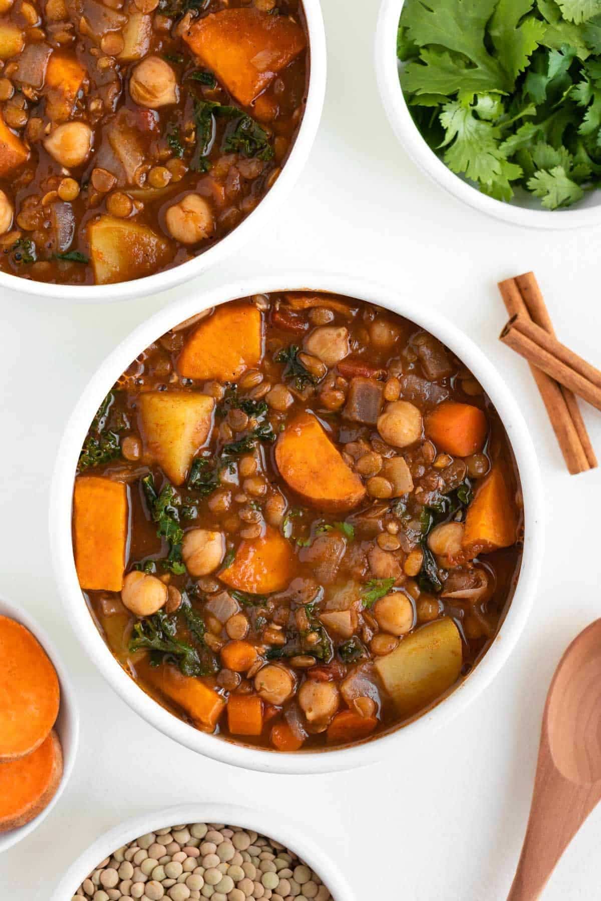 2 white bowls full of moroccan stew, with bowls on the side filled with clantro, cinnamon sticks and lentils