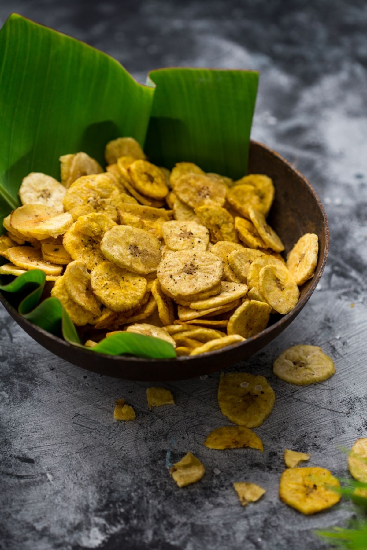 a bowl lined with a banan leaf filled with banana chips. Some have fallen onto the gray counter