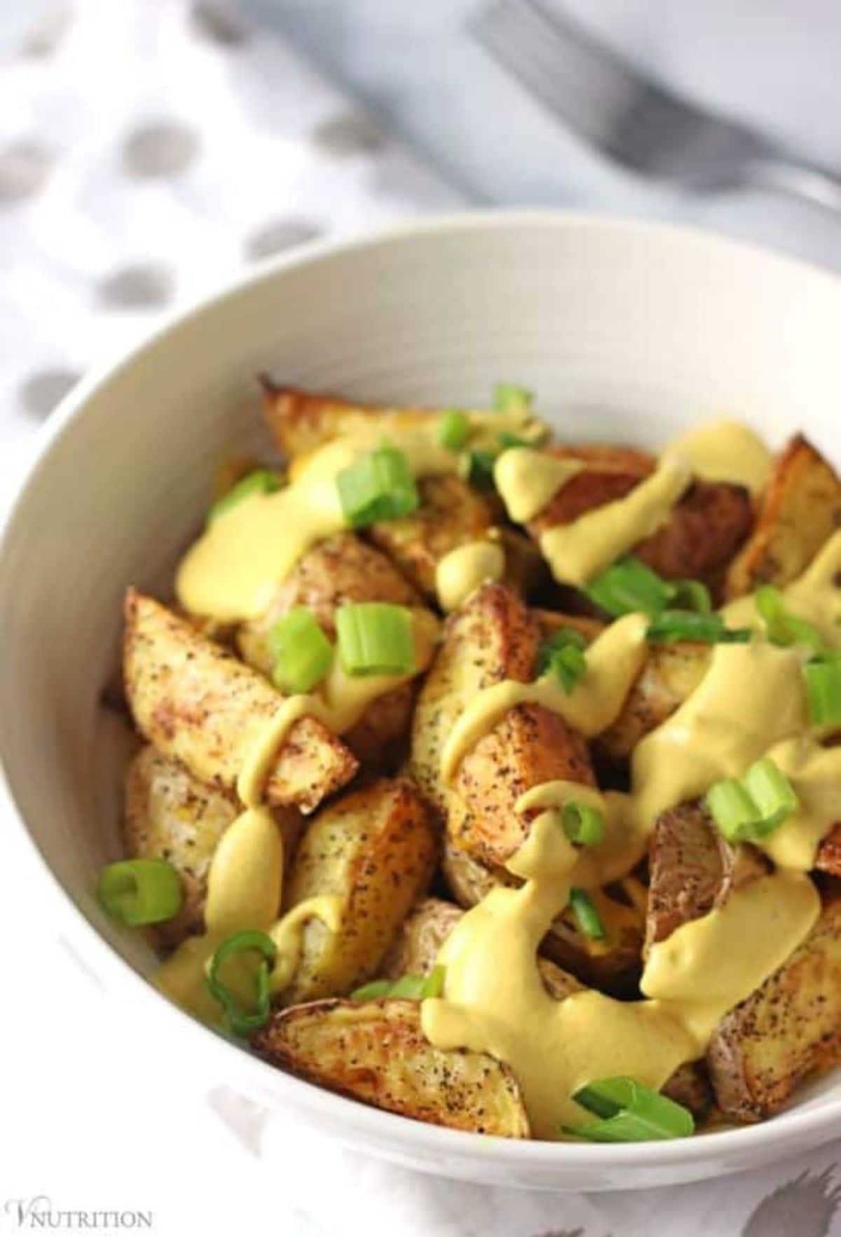 a white bowl full of potato wedges drizzled with yellow sauce and chopped scallions