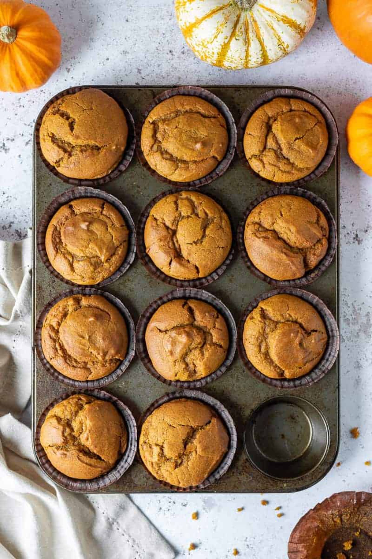 a muffin tray filled with pumpkin muffins. one is missing. small pumpkins sit around the tray