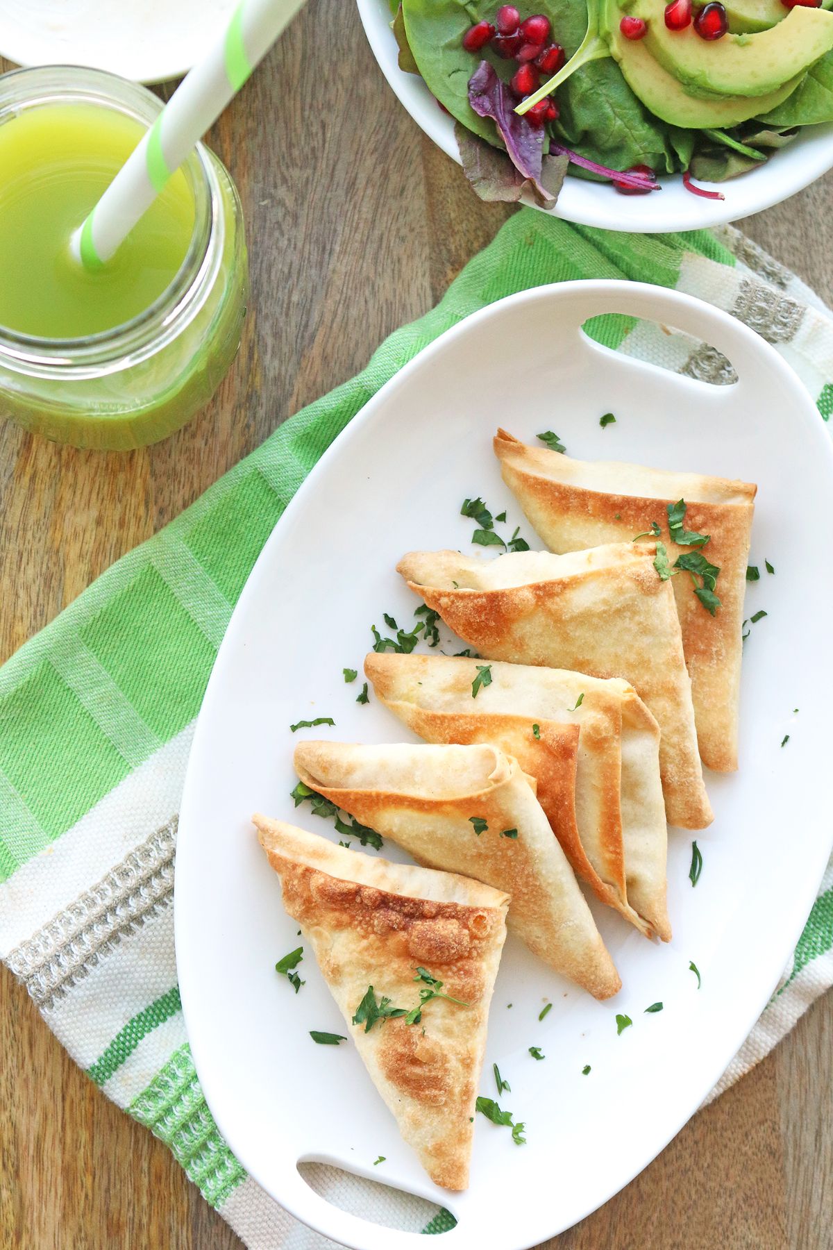 an oval dish with vegan samosas scattered with chopped herbs. The dish is surrounded by a bowl of salad and a glass full of green juice