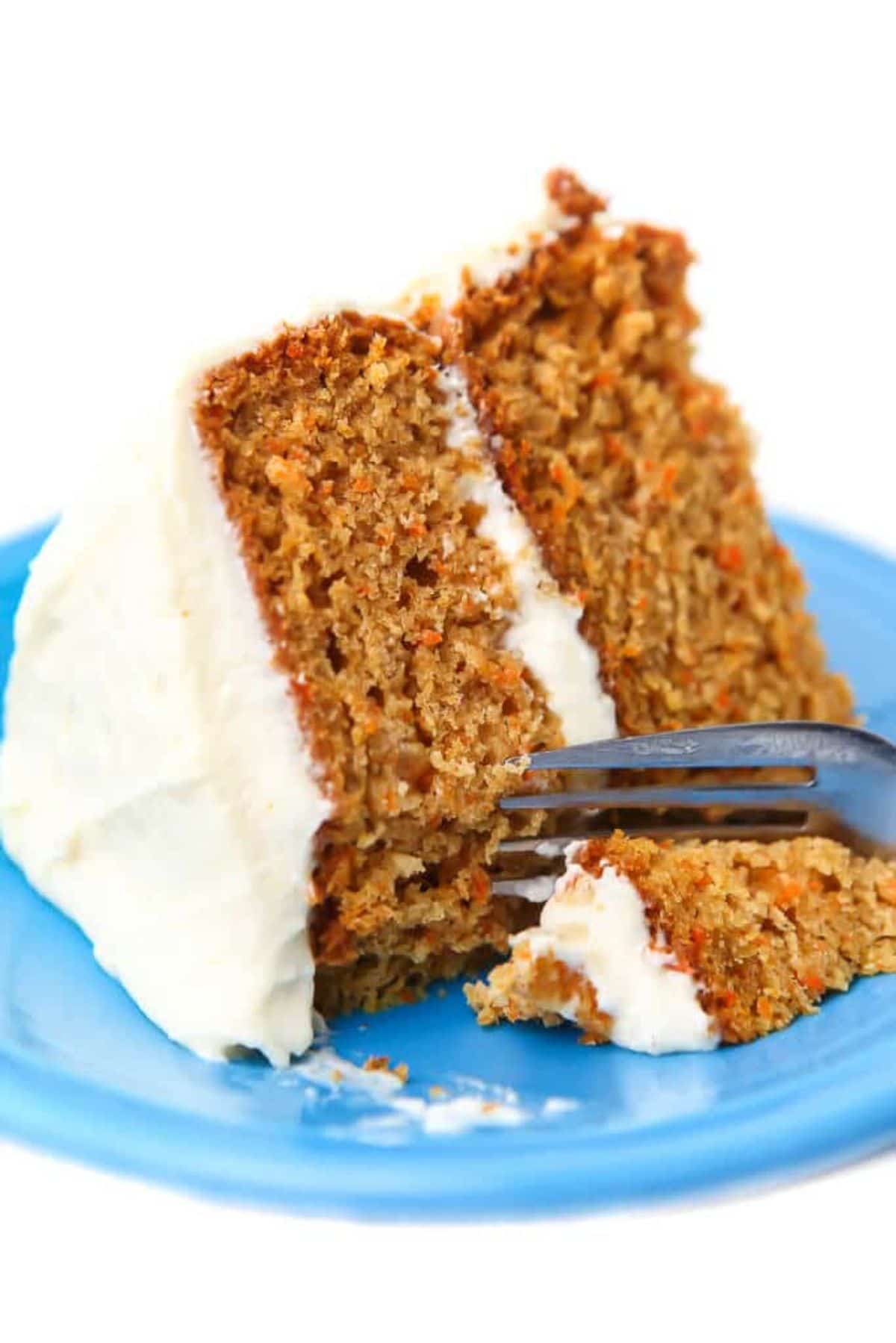 on a blue plate is a slice of carrot cake, iced, on it's side. A fork is cutting a seciton out of it