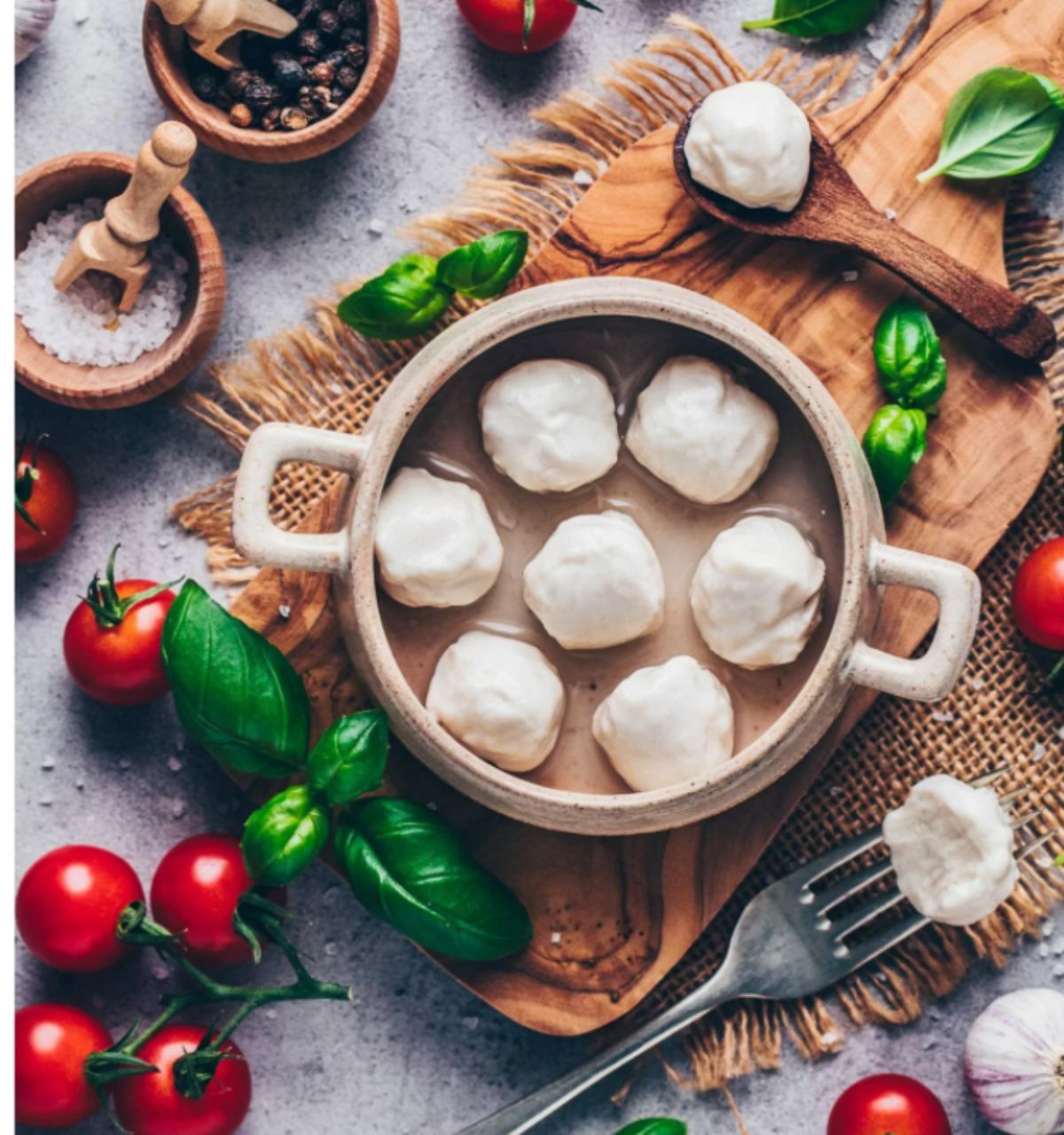 A dish of vegan mozzarella balls surrounded by fresh basil, cherry tomatoes and dishes of salt and pepper