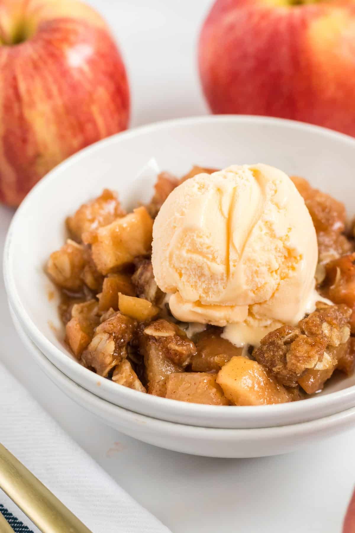 a bowl of apple crisp topped with vegan ice cream. 2 apples sit behind
