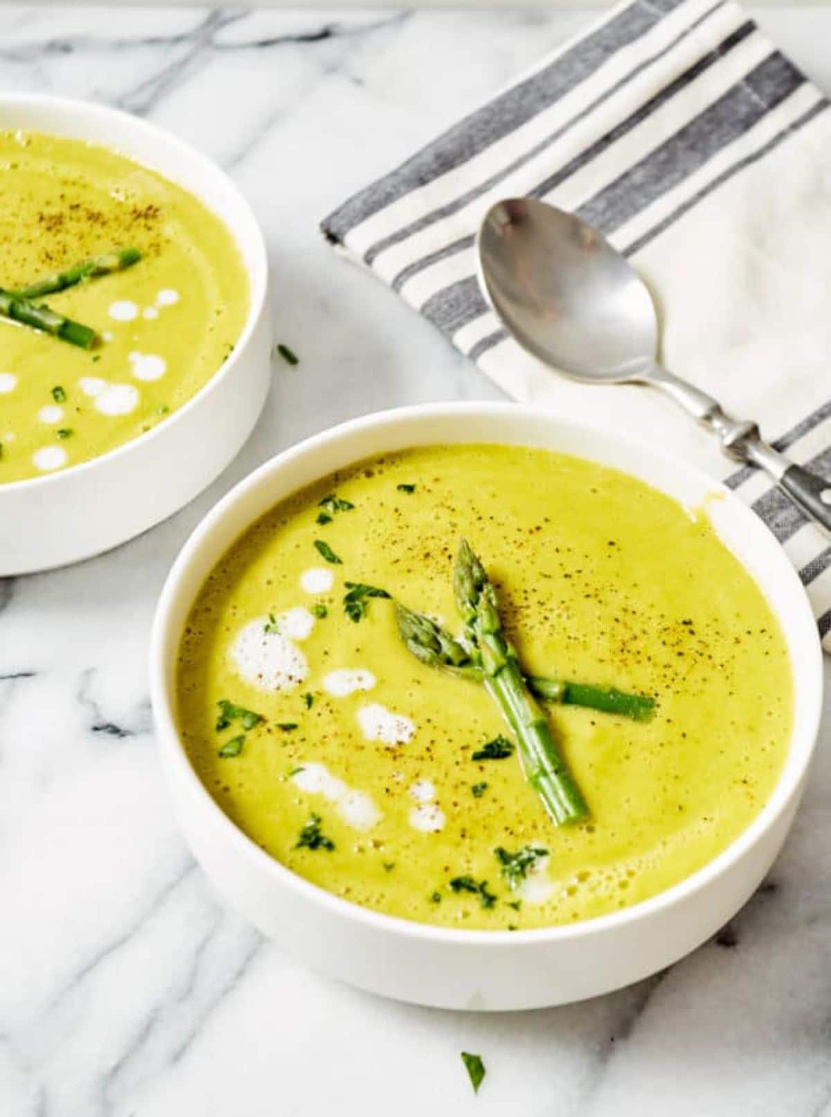 2 white bowls full of yellow asparagus soup, topped with spears of asparagus and herbs